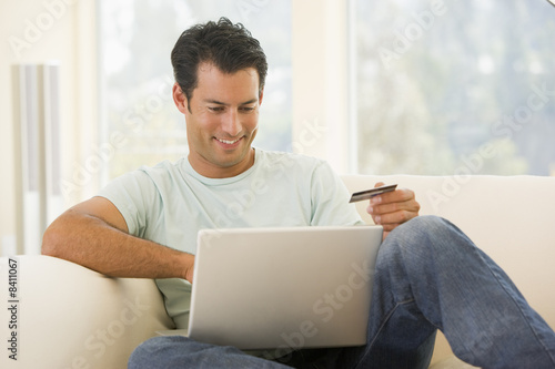 Photo: Man in living room using laptop and holding credit card smiling Man in living room using laptop and holding credit card smiling