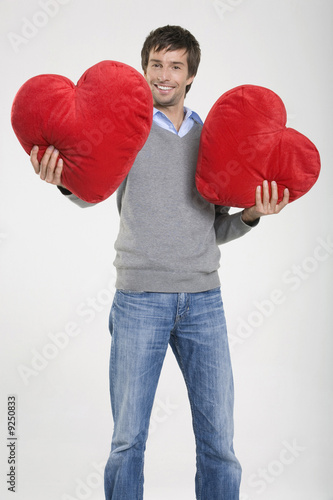 Photo: Young man holding heart-shaped cushions, portrait Young man holding heart-shaped cushions, portrait