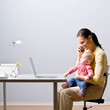 Working mother holding baby while talking on cell phone at desk