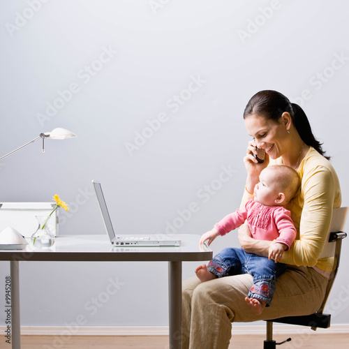 Photo: Working mother holding baby while talking on cell phone at desk Working mother holding baby while talking on cell phone at desk