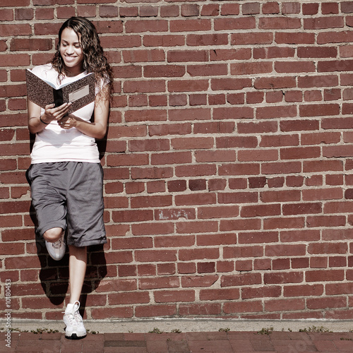 Photo: Beautiful young girl reading notebook against a brick wall. Beautiful young girl reading notebook against a brick wall.