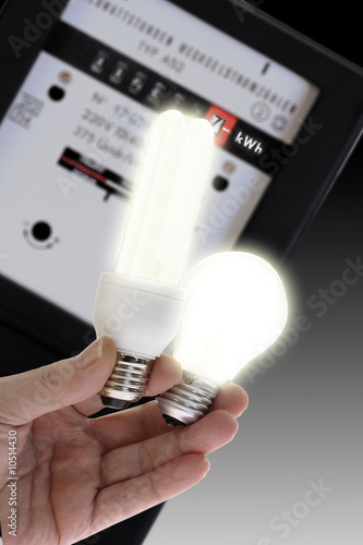 Photo: Energy saving lamp and electric bulb, electric meter in background, close-up Energy saving lamp and electric bulb, electric meter in background, close-up
