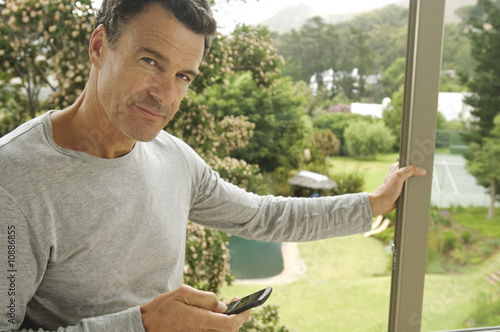 Photo: Man leaning against bay window, holding mobile phone Man leaning against bay window, holding mobile phone