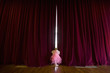 Ballerina girl (4-6) with fairy wings peeking out gap in stage curtains, rear view