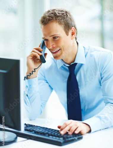 Foto: Young busineass man speaking on the phone while working on a com Young busineass man speaking on the phone while working on a com