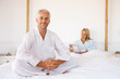 A senior man on bed with his wife in the background