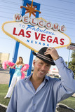 Photo: Portrait of mid-adult man in front of Welcome to Las Vegas sign, mid-adult woman in background. Portrait of mid-adult man in front of Welcome to Las Vegas sign, mid-adult woman in background.