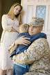 Military father embracing son 5-6 outside home, wife watching