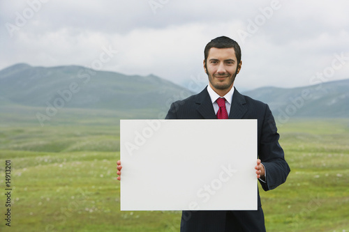 Photo: Smiling businessmen standing in mountain field holding blank sign, front view Smiling businessmen standing in mountain field holding blank sign, front view