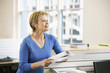 Photo: Middle-aged female office worker leaning on cubicle in office holding paperwork Middle-aged female office worker leaning on cubicle in office holding paperwork