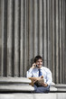 Businessman with newspaper sitting on pillar outside building talking on cell phone