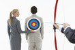 Photo: Business woman holding target to man's back while other man close-up of hands aims bow and arrow, against white background Business woman holding target to man's back while other man close-up of hands aims bow and arrow, against white background