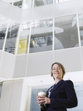 Photo: Business woman holding cup of coffee, standing in atrium of office building, low angle view Business woman holding cup of coffee, standing in atrium of office building, low angle view