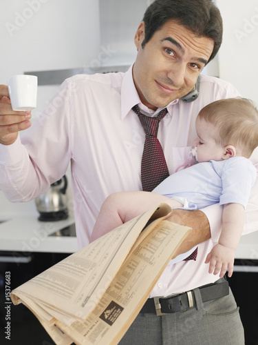 Photo: Father holding baby daughter while talking on phone in kitchen Father holding baby daughter while talking on phone in kitchen