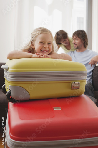 Photo: Germany, Leipzig, Girl (4-5) resting chin on suitcase, parents in background, smiling, portrait Germany, Leipzig, Girl (4-5) resting chin on suitcase, parents in background, smiling, portrait
