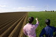 Photo: Farmers standing in young wheat field next to ploughed field and pointing at wind turbine in distance Farmers standing in young wheat field next to ploughed field and pointing at wind turbine in distance