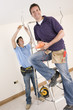 Smiling electrician holding voltmeter on ladder with co-worker wiring ceiling in background