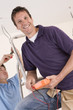 Smiling electrician holding voltmeter on ladder with co-worker wiring ceiling in background