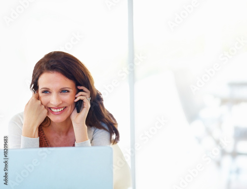 Photo: A cheerful woman using cellphone with laptop in front A cheerful woman using cellphone with laptop in front