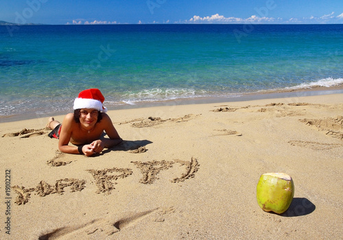 Photo: Young man in Santa hat on beach, with words happy on sand Young man in Santa hat on beach, with words happy on sand