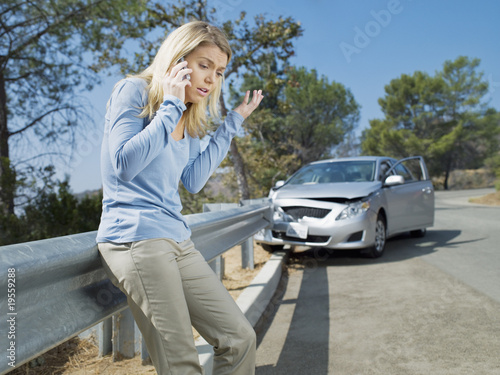 Foto: Frustrated woman using cell phone next to car wrecked on guardrail Frustrated woman using cell phone next to car wrecked on guardrail