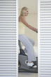 mature woman on exercise bike behind blinds pedalling side view