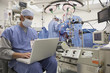 Doctor using laptop in operating room with surgery in background