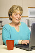 A senior woman works on her laptop in her kitchen, coffee mug at A senior woman works on her laptop in her kitchen, coffee mug at