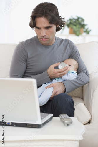 Photo: Hispanic father feeding baby and looking at laptop Hispanic father feeding baby and looking at laptop