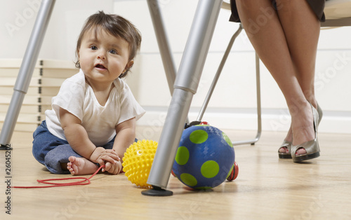 Photo: Hispanic baby boy sitting under mother's desk in home office Hispanic baby boy sitting under mother's desk in home office