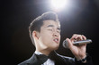 Asian man in tuxedo singing into microphone with eyes closed
