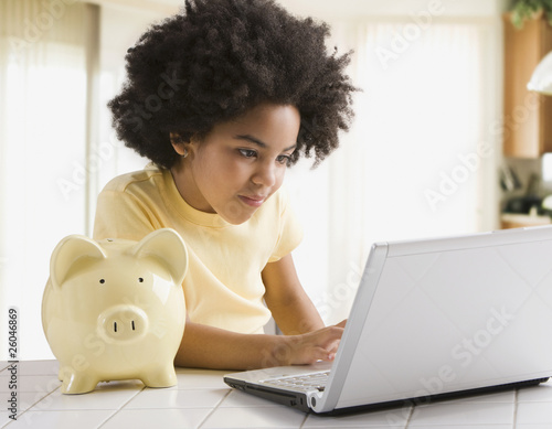 Photo: Mixed race girl using laptop next to piggy bank Mixed race girl using laptop next to piggy bank