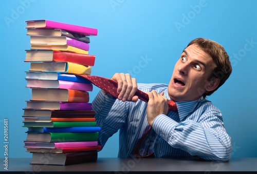 Photo: man at the table looking with fear at a stack of colored books man at the table looking with fear at a stack of colored books