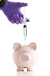 Gloved hand holding syringe over a piggy bank.