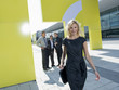 Photo: A blonde businesswoman outside a modern office building, three businessmen talking in the background A blonde businesswoman outside a modern office building, three businessmen talking in the background
