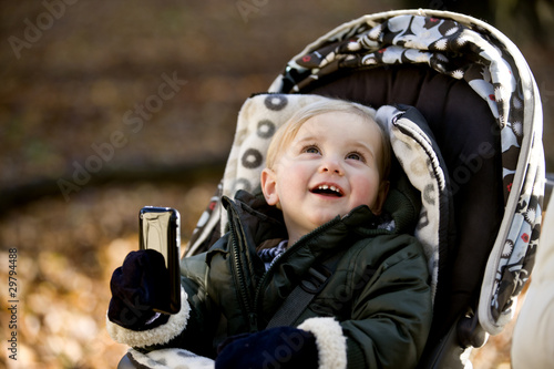 Photo: A young boy in a stroller holding an iphone, smiling A young boy in a stroller holding an iphone, smiling
