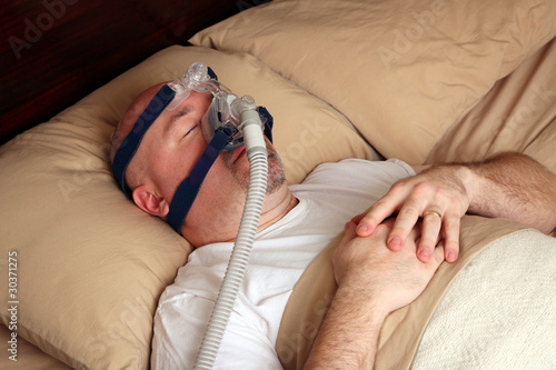 Photo: Man with sleep apnea using a CPAP machine in bed. Man with sleep apnea using a CPAP machine in bed.