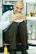 Man sitting in front of open refrigerator, eating hamburger