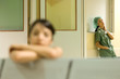 Photo: Medical worker standing in hospital waiting room, hands in pockets, looking at camera, woman resting head on arms in foreground Medical worker standing in hospital waiting room, hands in pockets, looking at camera, woman resting head on arms in foreground