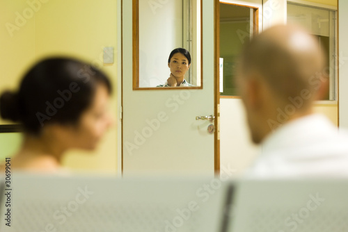 Photo: Medical worker looking through window, adults sitting in hospital waiting room in foreground Medical worker looking through window, adults sitting in hospital waiting room in foreground