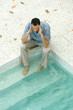 Man sitting with feet in pool, holding head, high angle view