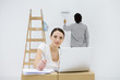 Young woman sitting at makeshift desk, using laptop computer, smiling at camera, man painting wall in background