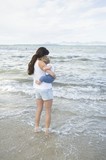 Photo: A lady holding a toddler with her feet in the shallow water, standing in front of the sea with mountains in the far background A lady holding a toddler with her feet in the shallow water, standing in front of the sea with mountains in the far background