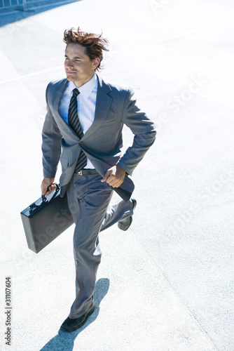 Photo: Young businessman running outdoors, carrying briefcase, smiling Young businessman running outdoors, carrying briefcase, smiling