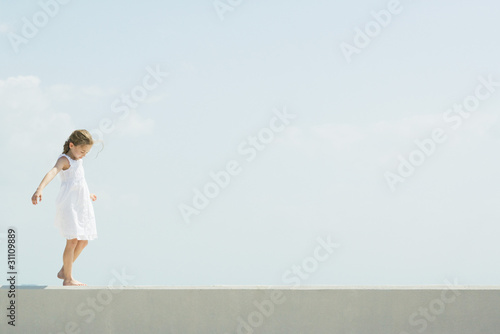 Photo: Little girl walking across low wall, arms out, looking down Little girl walking across low wall, arms out, looking down