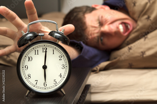 Photo: Alarm clock with male model in bed in background. Alarm clock with male model in bed in background.
