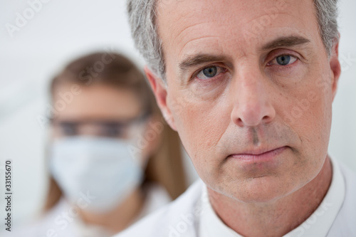 Photo: Serious man in lab coat and woman with surgical mask in background Serious man in lab coat and woman with surgical mask in background