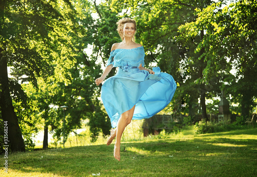 Photo: Young charming laughing woman in beautiful dress jumps on meadow Young charming laughing woman in beautiful dress jumps on meadow