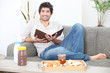 Man sitting on a sofa eating breakfast and reading a book