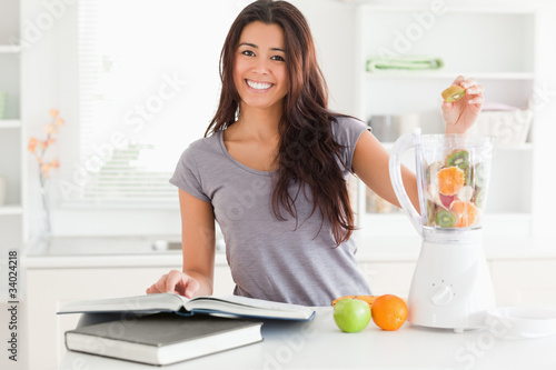 Photo: Beautiful woman consulting a notebook while filling a blender wi Beautiful woman consulting a notebook while filling a blender wi